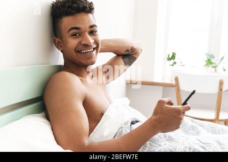 Image of optimistic pleased happy young african guy in the morning in bedroom using mobile phone in bed. Stock Photo