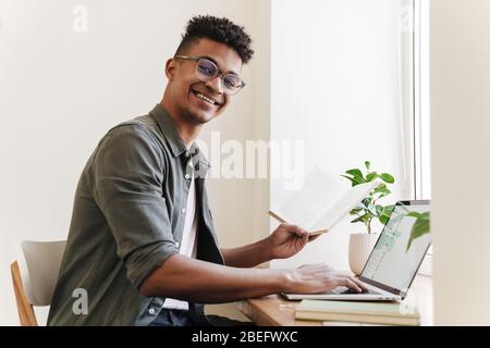 Picture of young happy positive smiling african guy in the morning using laptop computer holding notebook. Stock Photo