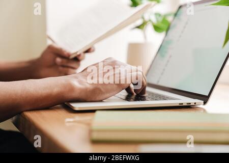 Cropped photo of young african guy indoors using laptop computer and holding notebook. Stock Photo
