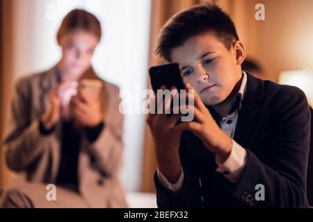 A kid sitting and playing on his smartphone, his older sister in the back also staring at her phone. Stock Photo