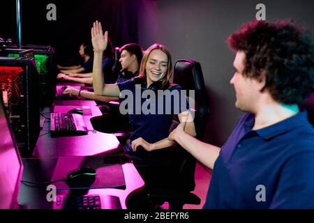Positive young woman with headphones around neck giving five to video game ally while celebrating success with him in computer club Stock Photo
