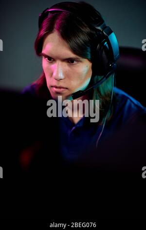 Focused guy with long hair using headset with microphone for communication with network gamers while playing computer game Stock Photo