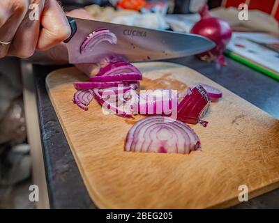 Human hand slicing red onions (Allium cepa) with a sharp kitchen knife on a wooden cutting board with selective focus on the sliced onions, i.e. fo Stock Photo