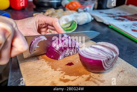 Human hand slicing red onion (Allium cepa) with a sharp kitchen knife on a wooden cutting board whilst preparing dinner. Selective and sharp focus Stock Photo