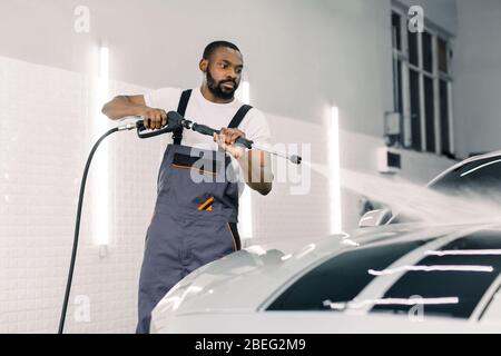 Shot of young black man in working clothes, car wash employee, washing the car hood under high pressure water gun at professional auto washing service Stock Photo
