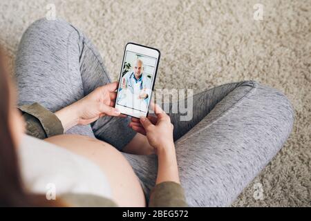 Above view of pregnant woman sitting with crossed legs on floor and using smartphone while talking to gynecologist via video chat Stock Photo