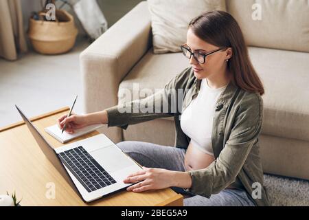 Content young pregnant woman in eyeglasses sitting at coffee table and using laptop while making notes about maternity at home Stock Photo