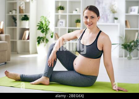 Portrait of smiling attractive young pregnant woman in sports bra and leggings sitting on yoga mat and twisting body aside Stock Photo