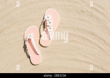 Top view of 2 white woman flip flops with flowers on golden sand beach near the sea. Summer vacation and relaxation concept. Copy space for text. Summ Stock Photo