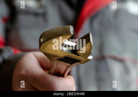 A man holds out an adjustable wrench into the camera. Mechanic in a red-gray working uniform with a tool in his hand. Close-up. Selective focus. Blurr Stock Photo