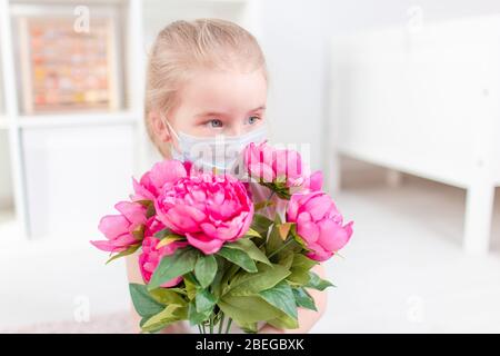 Little blonde girl with pollen allergy wearing medical mask and holding flowers. Girl smells flowers Stock Photo