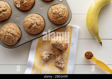 Banana Nut Muffins with One Torn Apart and Banana, Cinnamon and Kitchen Towel on White Wood Background Stock Photo