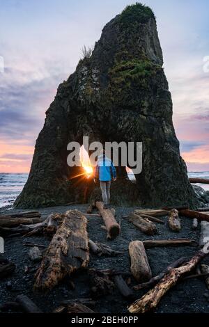 Man walking on log at the beach during sunset near dramatic rock Stock Photo