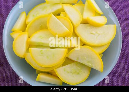 Sliced raw yellow summer crookneck squash in blue bowl on purple tablecloth Stock Photo