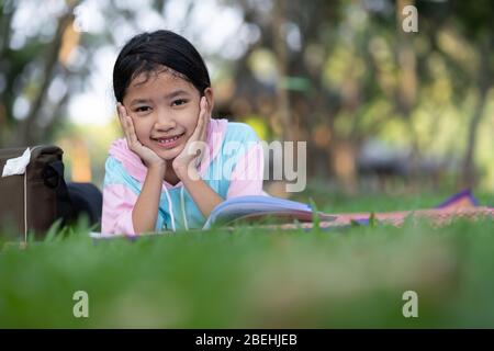 A little girl is reading a book under a tree on the grass in the garden with sunlight. Stock Photo