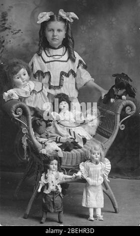About 1910, a Girl stands behind her family of dolls, which are poised on a wicker seat that has no back. The girl is herself very well dressed in a fancy dress and two bows in her long curled hair. Her family consists of 5 bisque dolls of different sizes and one Steiff Bear dressed in a large black bow. The large seated doll has a stuffed dog on a lease. The dog rests on the chair arm. This somewhat dangerous-looking dog, besides being collared, also has a large black bow around it's neck.   To see my other toy-related vintage images, Search:  Prestor  vintage  toy  [or kids] Stock Photo