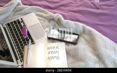Work from home, text written on notepad. Computer laptop, cellphone and notepad on top of bed in the bedroom. Stock Photo