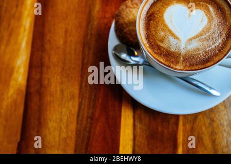 Close up cup of cappuccino in a white cup on a saucer with cookies. Top view of a drawing of a heart on coffee foam. Morning coffee stands on a wooden Stock Photo