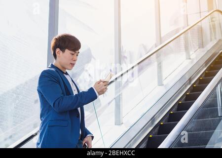 Asian handsome young business smiling and holding smartphone while standing on the escalator. Business and finance concept. Stock Photo