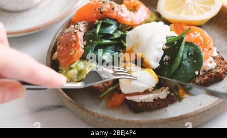 Eating toast with poached egg, salmon, avocado and spinach. Healthy lunch, breakfast or light dinner meal. Cutting poached egg with fork and knife Stock Photo