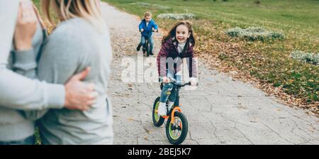 Cute girl with small brother riding the bike in the park while parents are watching them Stock Photo