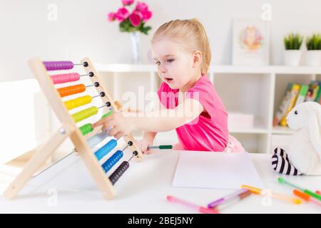 Little smiling blond girl sitting at the white desk and counting on the colourful abacus in the classroom. Preschool education Stock Photo