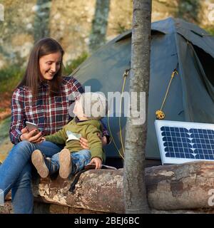 Woman looking at a child, smiling, sitting on a log in the forest near small solar panel and a tent camp. Girl is holding a telephone. Family weekend with the development of new technologies Stock Photo