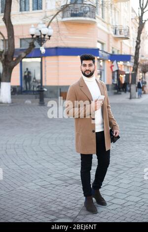 Portrait of fashionable well dressed bearded man at street. Young Indian man using app in his smartphone to take a taxi. Stock Photo