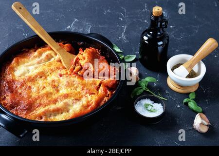 Italian cannelloni pasta stuffed with bolognese sauce and mozzarella cheese on top on a black baking dish on dark concrete background with fresh basil Stock Photo