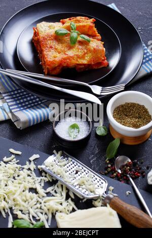 Italian pasta manicotti baked with beef bolognese sauce and mozzarella cheese on top on a black plate on dark concrete background with ingredients: ch Stock Photo