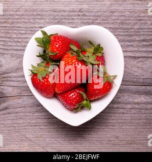 in the heart-shaped bowl of white ceramic, ripe strawberries, in the foreground, against the background of rough wood Stock Photo