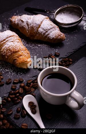cup of black coffee in the foreground and croissants with veiled sugar on a dark background Stock Photo