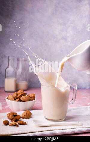 from the jug the almond milk is poured into the glass. on the table, bowl with almonds in shell. on the gray background some glass bottles. Stock Photo