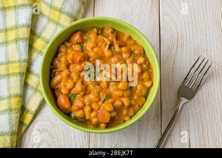 Backed beans with sausage slices in tomato sauce and parsley in a green bowl and fork on a wood table. Bean is source of vegetable protein. Stock Photo