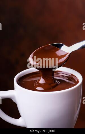 in the foreground, hot and liquid chocolate pouring from the spoon into a white ceramic cup Stock Photo