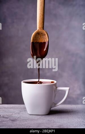 in the foreground, hot and liquid chocolate pouring from the spoon into a white ceramic cup Stock Photo