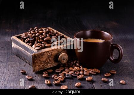 On the raw and dark wooden table, a cup of hot coffee and a drawer of an old grinder with roasted coffee beans. Still life Stock Photo
