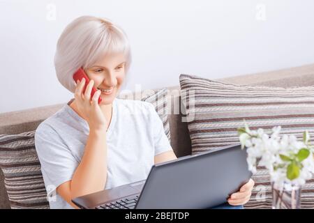 Pretty young smiling woman is sitting on the sofa at home and holding a laptop while talking on the phone. Work from home, freelance concept Stock Photo