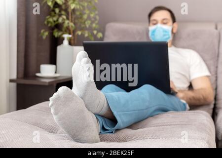 Young caucasian man working from home wearing protective mask using laptop and internet. Cozy Home office, workplace on sofa during coronavirus Stock Photo