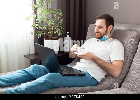 Happy young caucasian man in medical face mask drink tea while working from home using laptop. Cozy home office, workplace on sofa on coronavirus Stock Photo