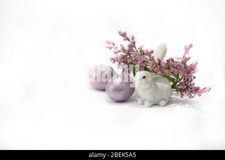 A white bunny rabbit with wreath of purple flowers and two colored easter eggs near. Easter holiday concept. Stock Photo