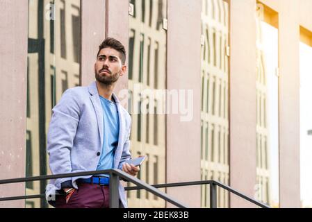 Portrait of a happy bearded young man holding a smartphone while standing outdoors and looking away Stock Photo