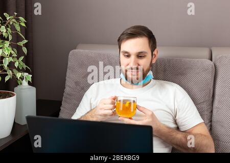 Happy young caucasian man in medical face mask drink tea while working from home using laptop. Cozy home office, workplace on sofa on coronavirus Stock Photo