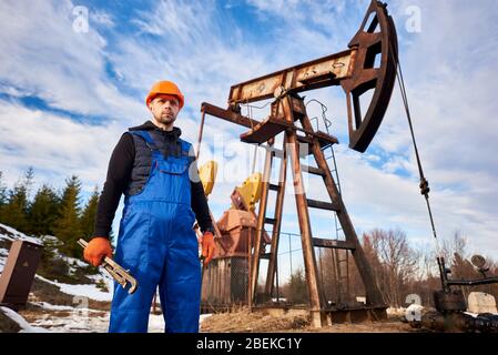 Portrait of oil well operator in protective helmet and work overalls holding industrial wrench. Male worker standing near petroleum pump jack under beautiful sky. Concept of petroleum industry. Stock Photo