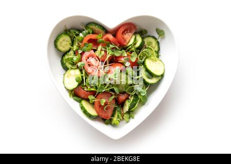 Salad with tomato, cucumber, vegetables, microgreen radishes in plate shape of heart. Isolated on white. Concept vegan and healthy eating. Top view. Stock Photo
