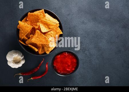 Mexican nachos in a black bowl with hot salsa dip sauce, garlic and chili peppers on textured black background. Top view with copy space. Stock Photo