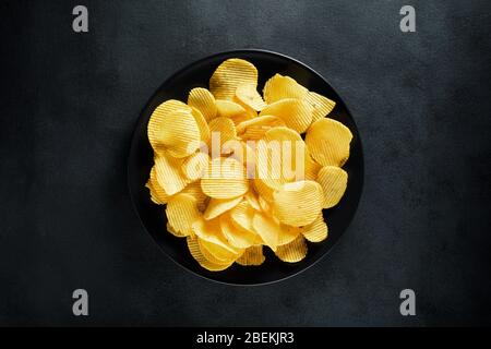 Crispy potato chips on a black plate with black textured background. Top view. Stock Photo