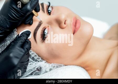 Professional cosmetologist hands are doing permanent makeup on eyebrows of young woman, close up. Stock Photo