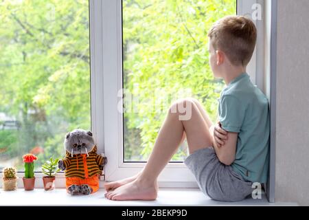 Alone sadness caucasian boy sitting on windowsill, looking outdoors during pandemic of coronavirus covid-19. Feels unhappy and lonely. Stay at home Stock Photo