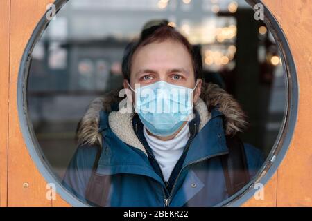 Man looks out the round window, wears protective medical mask against transmissible infectious diseases and as protection against flu or coronavirus, Stock Photo